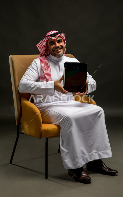 Displaying applications and services for the tablet device, a portrait of a smiling Saudi Arabian Gulf man wearing a shemagh and traditional thobe, sitting on a chair holding a tablet with a blank black screen in his hands, using advanced technology for electronic digital marketing, expressions of joy and happiness, black background