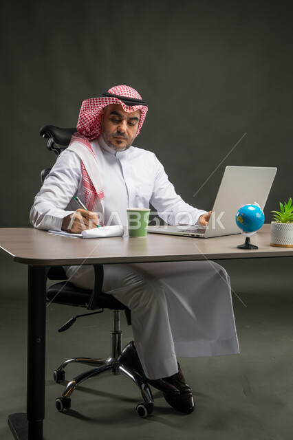Attending online training workshops for development in the practical field, holding a business meeting online via the laptop, a portrait of a smiling Saudi Arabian Gulf man wearing a shemagh and traditional thobe, sitting at the desk completing work tasks and taking notes and data, black background.
