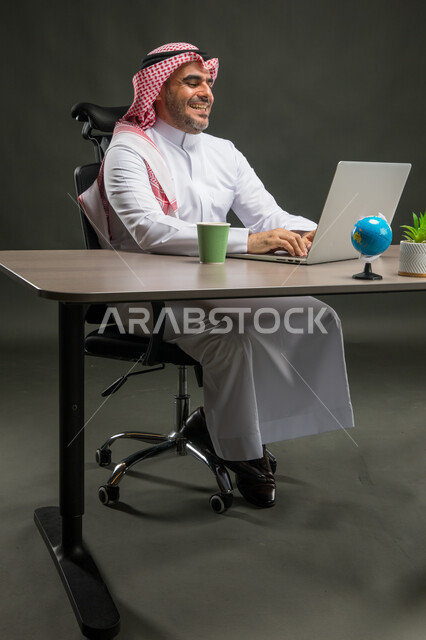 مهن ووظائف مكتبية سعودية، مفهوم ادارة الاعمال وتحقيق اهداف الشركة، بورتريه لرجل اعمال عربي خليجي سعودي يرتدي الشماغ والثوب التقليدي يجلس في مكتبه يعمل على الحاسوب المحمول بايماءات الحماس والسعادة، انجاز مهام العمل عن بعد، خلفية سوداء