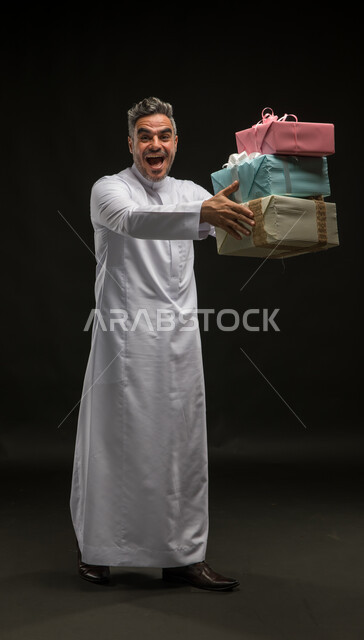 Expressions of enthusiasm and joy for holidays, happy occasions and surprises, a portrait of a smiling Saudi Arabian Gulf man wearing a traditional dress, holding wrapped gift boxes in his hands, looking at the camera, expressing love and familiarity between friends, offering gifts and congratulations, full body portrait, black background