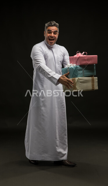 إيماءات الحماس والسرور، أعياد ومناسبات ومفاجآت سعيدة, بورتريه لرجل عربي خليجي سعودي مبتسم يرتدي الثوب التقليدي يحمل بيديه علب هدايا مغلفة ينظر الى الكاميرا، التعبير عن المحبة والألفة بين الأصدقاء, الحصول على الهدايا والتهنئات، صورة الجسم بالكامل، خلفية سوداء