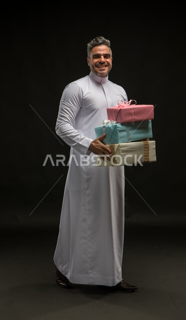 أعياد ومناسبات ومفاجآت سعيدة, بورتريه لرجل عربي خليجي سعودي مبتسم يرتدي الثوب التقليدي يحمل بيديه علب هدايا مغلفة ينظر الى الكاميرا بإيماءات الخجل والسرور، التعبير عن المحبة والألفة بين الأصدقاء, الحصول على الهدايا والتهنئات، صورة الجسم بالكامل، خلفية سوداء