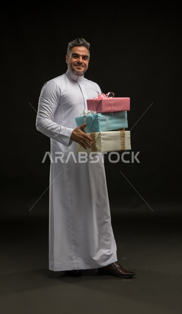 الحصول على الهدايا والتهنئات، أعياد ومناسبات ومفاجآت سعيدة, بورتريه لرجل عربي خليجي سعودي مبتسم يرتدي الثوب التقليدي يحمل بيديه علب هدايا مغلفة ينظر الى الكاميرا بإيماءات الخجل والسرور، التعبير عن المحبة والألفة بين الأصدقاء, صورة الجسم بالكامل، خلفية سوداء