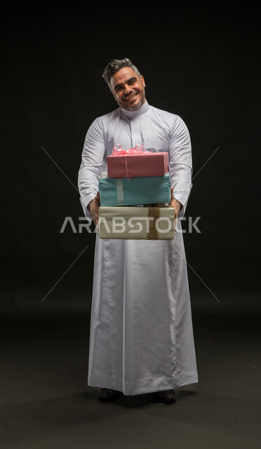 التعبير عن المحبة والألفة بين الأصدقاء, تقديم الهدايا والتهنئات، أعياد ومناسبات ومفاجآت سعيدة, بورتريه لرجل عربي خليجي سعودي مبتسم يرتدي الثوب التقليدي يحمل بيديه علب هدايا مغلفة ينظر الى الكاميرا بإيماءات الخجل والسرور، صورة الجسم بالطول الكامل، خلفية سوداء