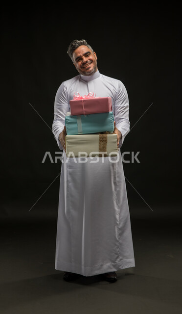 تقديم الهدايا والتهنئات، أعياد ومناسبات ومفاجآت سعيدة, بورتريه لرجل عربي خليجي سعودي مبتسم يرتدي الثوب التقليدي يحمل بيديه علب هدايا مغلفة ينظر الى الكاميرا بإيماءات الخجل والسرور، التعبير عن المحبة والألفة بين الأصدقاء, صورة الجسم بالطول الكامل، خلفية سوداء