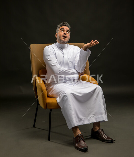 تعابير الصدمة والتعجب من شيء ما، النظر إلى عنصرما عاليا، الاهتمام بالمظهر الخارجي، بورتريه لرجل عربي خليجي سعودي مبتسم يرتدي الثوب التقليدي يرفع كفه للأعلى بإيماءات الاستغراب والدهشة، خلفية سوداء.