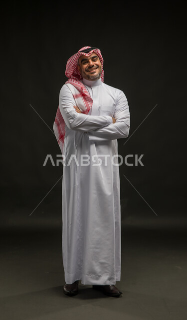 مفهوم الرجولة والأناقة، بورتريه لرجل عربي خليجي سعودي مبتسم يرتدي الشماغ والثوب التقليدي يقف بإستقامة وأيدي متقاطعة ينظر إلى الكاميرا بإيماءات البشاشة والسرور، الاهتمام بالمظهر الخارجي، صورة الجسم بالطول الكامل، خلفية سوداء