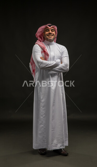 Concern with external appearance, the concept of masculinity and elegance, a portrait of a smiling Saudi Arabian Gulf man wearing a shemagh and traditional dress, standing straight with crossed hands looking at the camera with gestures of joy and pleasure, full-length body portrait, black background