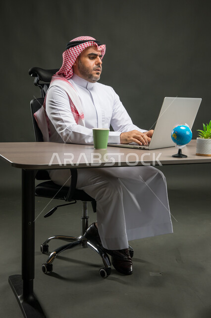 إيماءات الجدية والحزم، الشعور بالانزعاج والتعب خلال فترة العمل، بورتريه لرجل اعمال عربي خليجي سعودي يرتدي الشماغ والثوب التقليدي يجلس في مكتبه يستخدم الحاسوب المحمول، إنجاز مهام العمل اليومية، خلفية سوداء