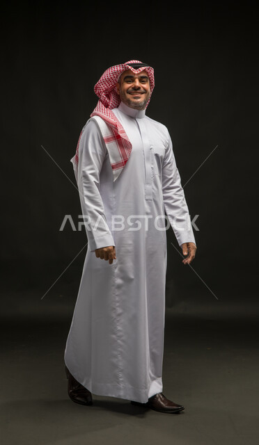 Walking and going somewhere, the concept of caring about external appearance and elegance, portrait of a smiling Saudi Arabian Gulf man wearing a shemagh and traditional dress looking at the camera with gestures of joy and happiness, full-length body portrait, black background