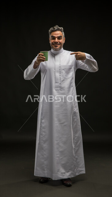تناول المشروبات الساخنة، الشعور بالانتعاش والفرح، الاستمتاع بمذاق القهوة الزاكي، الحيوية والنشاط عند شرب الكافيين، بورتريه لرجل عربي سعودي خليجي مبتسم يرتدي الثوب التقليدي يحمل بيده كوب ورقي ويشير إليه، صورة الجسم بالطول الكامل، خلفية سوداء