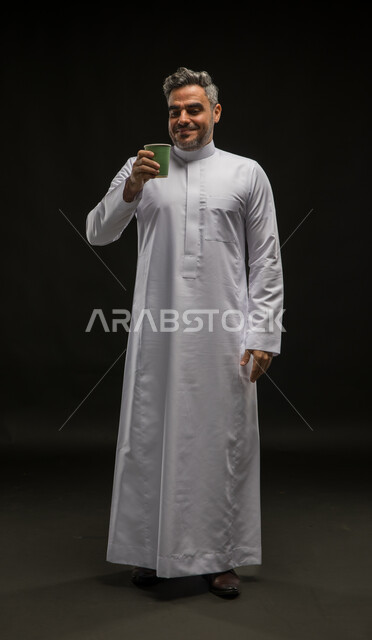 Feeling refreshed and happy, enjoying the good taste of coffee, vitality and activity when drinking caffeine, drinking hot drinks, portrait of a smiling Saudi Arabian Gulf man wearing a traditional dress holding a paper cup in his hand, full-length body portrait, black background