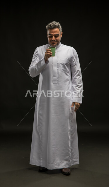احتساء المشروبات الساخنه والأعشاب، الاستمتاع بتناول القهوة العربية، تعديل المزاج بتناول الكافيين، بورتريه لرجل عربي خليجي سعودي مبتسم يرتدي الثوب التقليدي يحمل بيده كوب من الورق وينظر اليه, صورة الجسم بالطول الكامل، خلفية سوداء