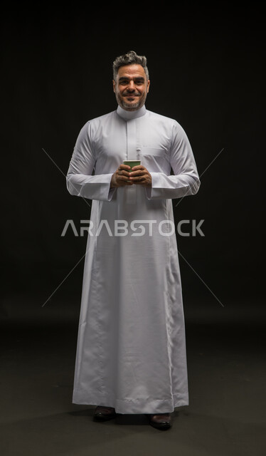 الشعور والاحساس بالدفء عند تناول المشروبات الساخنة، الاستمتاع بمذاق القهوة الزاكي، الحيوية والنشاط عند شرب الكافيين، بورتريه لرجل عربي سعودي خليجي مبتسم يرتدي الثوب التقليدي يحمل بيديه كوب ورقي، صورة الجسم بالطول الكامل، خلفية سوداء