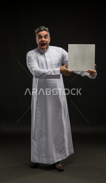 عرض لوحه بيضاء مربعه فارغة بإيماءات الاندهاش والاستغراب، استخدام لوحات توضيحية تعريفية، إعلانات وعروضات تسويقية، بورتريه لرجل عربي خليجي سعودي يرتدي الثوب التقليدي يحمل بيده موك اب مربع أبيض ينظر إلى الكاميرا، صورة الجسم بالطول الكامل، خلفية سوداء