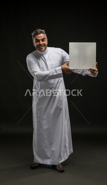استخدام لوحات توضيحية تعريفية، إعلانات وعروضات تسويقية، لوحه بيضاء مربعه فارغة، بورتريه لرجل عربي خليجي سعودي يرتدي الثوب التقليدي يحمل بيده موك اب مربع أبيض ينظر إلى الكاميرا، صورة الجسم بالطول الكامل، خلفية سوداء