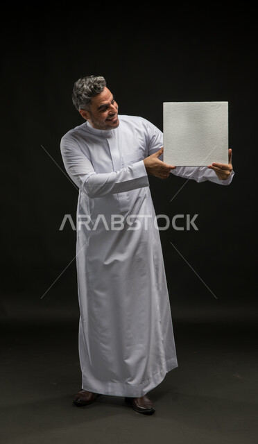 Full-length body portrait, white square mock-up, use of explanatory boards, portrait of a Saudi Gulf Arab man wearing a traditional dress, holding a blank square white board in his hand and looking at it with gestures of amazement and pleasure, advertisements and marketing presentations, black background