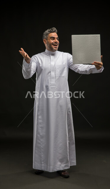 موك اب مربع أبيض، استخدام لوحات توضيحية تعريفية، بورتريه لرجل عربي خليجي سعودي يرتدي الثوب التقليدي يحمل بيده لوحه بيضاء مربعه فارغة وينظر اليها  بإيماءات الاندهاش والسرور، إعلانات وعروضات تسويقية، صورة الجسم بالطول الكامل، خلفية سوداء