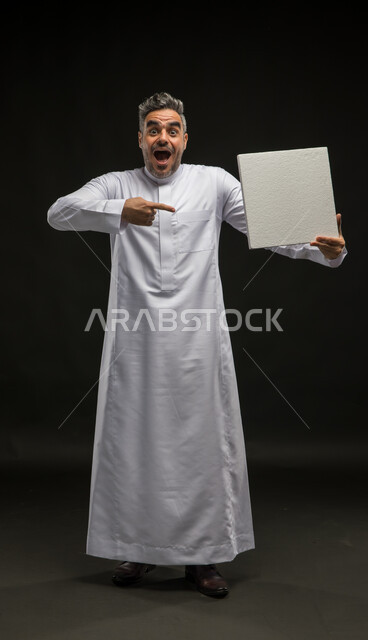 استخدام لوحات توضيحية تعريفية، الاشارة الى لوحه بيضاء مربعه فارغة بإيماءات الاندهاش والسرور، بورتريه لرجل عربي خليجي سعودي يرتدي الثوب التقليدي يحمل بيده موك اب مربع أبيض ينظر إلى الكاميرا، إعلانات وعروضات تسويقية، صورة الجسم بالطول الكامل، خلفية سوداء