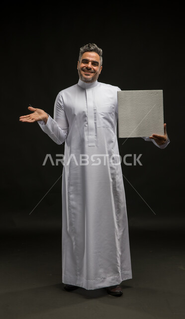 لوحه بيضاء مربعه فارغة، بورتريه لرجل عربي خليجي سعودي يرتدي الثوب التقليدي يحمل بيده موك اب مربع أبيض ينظر إلى الكاميرا، إعلانات وعروضات تسويقية، استخدام لوحات توضيحية تعريفية، صورة الجسم بالطول الكامل، خلفية سوداء