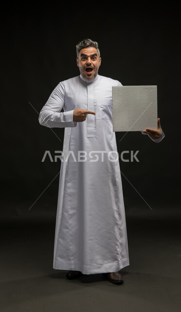 الاشارة الى لوحه بيضاء مربعه فارغة بإيماءات الاندهاش والسرور، بورتريه لرجل عربي خليجي سعودي يرتدي الثوب التقليدي يحمل بيده موك اب مربع أبيض ينظر إلى الكاميرا، إعلانات وعروضات تسويقية، استخدام لوحات توضيحية تعريفية، صورة الجسم بالطول الكامل، خلفية سوداء