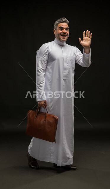 الترحيب والسلام برحابة صدر، بورتريه لرجل أعمال عربي خليجي سعودي مبتسم يرتدي الثوب التقليدي يحمل بيده حقيبة جلد بني يلوح بيده الى شخص ما، مفهوم إدارة الأعمال والمشاريع، صورة الجسم بالكامل، خلفية سوداء