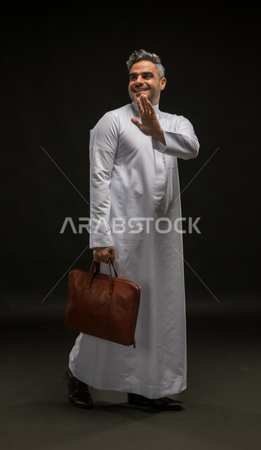 السلام والترحيب برحابة صدر، بورتريه لرجل أعمال عربي خليجي سعودي مبتسم يرتدي الثوب التقليدي يحمل بيده حقيبة جلد بني يلوح بيده الى شخص ما، مفهوم إدارة الأعمال والمشاريع، صورة الجسم بالكامل، خلفية سوداء