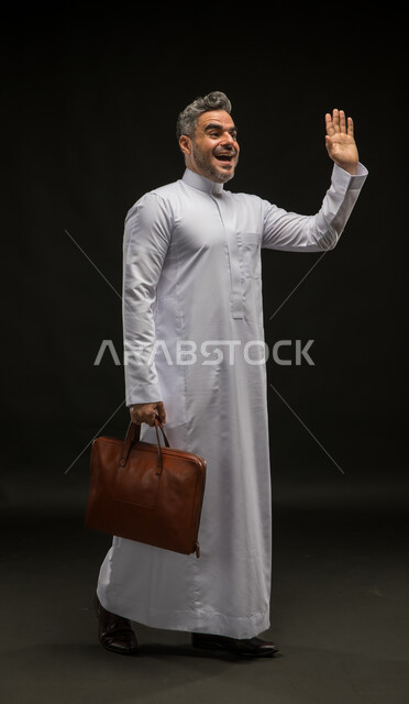 مفهوم إدارة الأعمال والمشاريع، السلام والترحيب برحابة صدر، بورتريه لرجل أعمال عربي خليجي سعودي مبتسم يرتدي الثوب التقليدي يحمل بيده حقيبة جلد بني يلوح بيده الى شخص ما، صورة الجسم بالكامل، خلفية سوداء