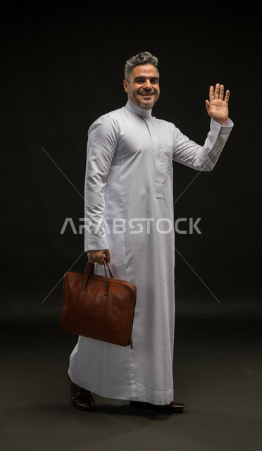 مفهوم إدارة الأعمال والمشاريع، السلام والترحيب برحابة صدر، بورتريه لرجل أعمال عربي خليجي سعودي مبتسم يرتدي الثوب التقليدي يحمل بيده حقيبة جلد بني يلوح بيده الى شخص ما، صورة الجسم بالكامل، خلفية سوداء