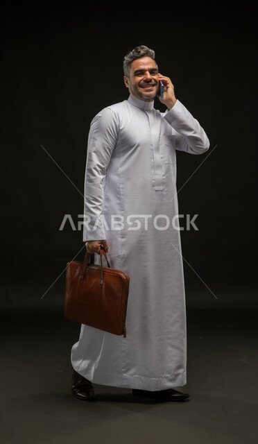 مفهوم إدارة الاعمال والمشاريع، بورتريه لرجل أعمال عربي خليجي سعودي مبتسم يرتدي الثوب التقليدي يحمل بيده حقيبة يد بني يقوم بإجراء اتصال هاتفي باستخدم الجوال ينظر الى الكاميرا، تحديد مواعيد اجتماعات العمل عبر الهاتف المحمول، صورة الجسم بالكامل، خلفية سوداء