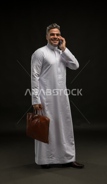تحديد مواعيد اجتماعات العمل عبر الهاتف المحمول، بورتريه لرجل أعمال عربي خليجي سعودي مبتسم يرتدي الثوب التقليدي يحمل بيده حقيبة يد بني يقوم بإجراء اتصال هاتفي باستخدم الجوال ينظر الى الكاميرا، مفهوم إدارة الاعمال والمشاريع، صورة الجسم بالكامل، خلفية سوداء