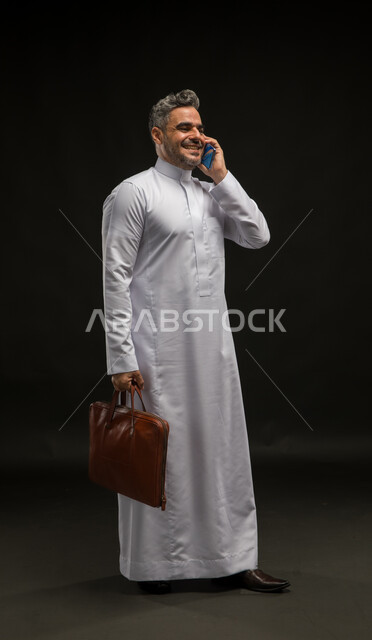 تحديد مواعيد اجتماعات العمل عبر الهاتف المحمول، بورتريه لرجل أعمال عربي خليجي سعودي مبتسم يرتدي الثوب التقليدي يحمل بيده حقيبة يد بني يقوم بإجراء اتصال هاتفي باستخدم الجوال، مفهوم إدارة الاعمال والمشاريع، صورة الجسم بالكامل، خلفية سوداء