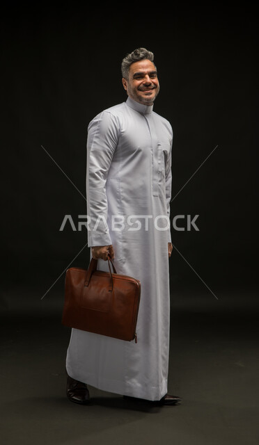 الاهتمام بالمظهر الخارجي والأناقة، بورتريه لرجل أعمال عربي خليجي سعودي مبتسم يرتدي الثوب التقليدي يحمل بيده حقيبة جلد بني ينظر إلى الكاميرا بإيماءات البشاشة والسرور، مفهوم ادارة الاعمال والمشاريع، المشي والذهاب الى مكان ما، صورة الجسم بالطول الكامل، خلفية سوداء