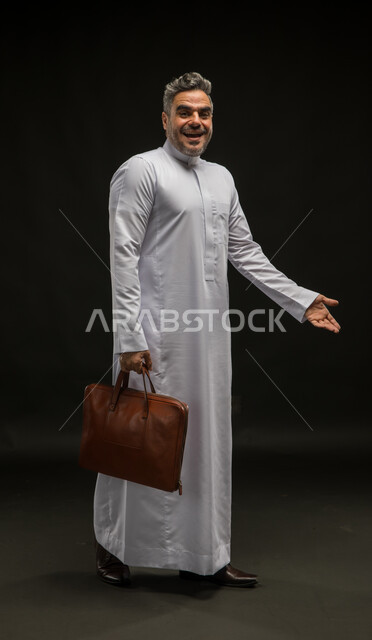 تعابير السعادة لرؤية شخص ما، مفهوم إدارة الأعمال والمشاريع، السلام والترحيب برحابة صدر، بورتريه لرجل أعمال عربي خليجي سعودي مبتسم يرتدي الثوب التقليدي يحمل بيده حقيبة جلد بني ينظر الى الكاميرا، صورة الجسم بالكامل، خلفية سوداء