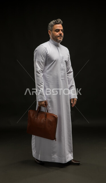 الأثر السلبي لضغوط العمل على صحة الانسان الجسدية والنفسية، الشعور بالتعب والارهاق، بورتريه لرجل أعمال عربي خليجي سعودي يرتدي الثوب التقليدي يحمل بيده حقيبة جلد بني بإيماءات اليأس والخمول، العودة من العمل بعد يوم شاق ومتعب، صورة الجسم بالكامل، خلفية سوداء