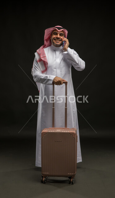 إجراء مكالمات هاتفية، الاستعداد لقضاء رحلة صيفية ممتعة, إيماءات السرور والسعادة, بورتريه لرجل عربي سعودي خليجي مبتسم يرتدي الشماغ والثوب التقليدي ينظر إلى الكاميرا يمسك بيده حقيبة السفر والهاتف المحمول، صورة للجسم بالكامل, خلفية سوداء.