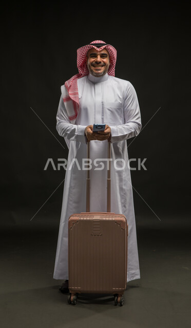 ايماءات النشاط والحيوية و الاستعداد لقضاء رحلة صيفية ممتعة, إيماءات السرور والسعادة, بورتريه لرجل عربي سعودي خليجي مبتسم يرتدي الشماغ والثوب التقليدي ينظر إلى الكاميرا يمسك بيده حقيبة السفر والهاتف المحمول، صورة للجسم بالكامل, خلفية سوداء.