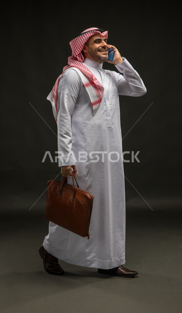 الشعور بالسرور لتحقيق أهداف الشركة, الذهاب إلى دوام العمل, بورتريه لرجل عربي سعودي خليجي مبتسم يرتدي الشماغ والثوب التقليدي يحمل حقيبة يد يقوم بإجراء مكالمة صوتية عبر الهاتف المحمول, الضحك والتسامر مع الأصدقاء، صورة الجسم بالكامل, خلفية سوداء