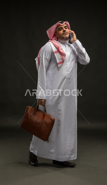 إيماءات الجدية والحزم, بورتريه لرجل عربي سعودي خليجي يرتدي الشماغ والثوب التقليدي يقف باستقامة يحمل حقيبة يد ويقوم بإجراء مكالمة صوتية عبر الهاتف المحمول, الذهاب إلى العمل في مقر الشركة، صورة الجسم بالكامل, خلفية سوداء