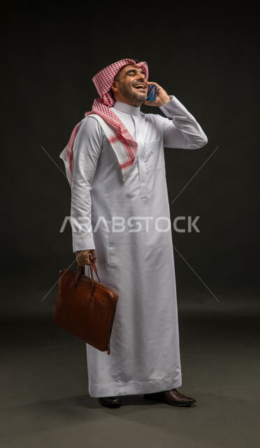 الذهاب إلى دوام العمل, الشعور بالسرور لتحقيق أهداف الشركة, بورتريه لرجل عربي سعودي خليجي مبتسم يرتدي الشماغ والثوب التقليدي يحمل حقيبة يد يقوم بإجراء مكالمة صوتية عبر الهاتف المحمول, الضحك والتسامر مع الأصدقاء، صورة الجسم بالكامل, خلفية سوداء