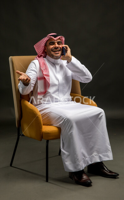 تبادل الاحاديث المسلية والحوارات، سماع الأخبار السارة والمفرحة، التسامر مع الأصدقاء والأقارب بإيماءات السعادة، بورتريه لرجل عربي خليجي سعودي مبتسم يرتدي الشماغ والثوب التقليدي يجلس على كرسي مريح يقوم بإجراء مكالمة صوتيه عبر الهاتف المحمول، خلفية سوداء