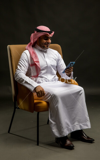 الدردشة والتسامر مع الأصدقاء والأقارب بإيماءات السعادة، سماع الأخبار السارة والمفرحة، بورتريه لرجل عربي خليجي سعودي مبتسم يرتدي الشماغ والثوب التقليدي يجلس على كرسي مريح يقوم بإجراء مكالمة صوتيه عبر الهاتف المحمول، خلفية سوداء