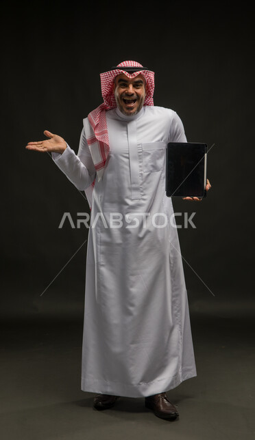 Displaying tablet applications and programs, the concept of remote project management, using modern technical devices, a portrait of a Saudi Gulf Arab man wearing a shemagh and traditional thobe, standing straight looking at the camera, showing the portable tablet with a blank black screen, full-length body portrait, black background