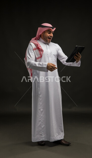 إيماءات وتعابير الفرح والسرور بتحقيق الأهداف, مفهوم إدارة المشاريع عن بعد, إستخدام اجهزة تقنية حديثة، بورتريه لرجل عربي خليجي سعودي يرتدي الشماغ والثوب التقليدي يقف باستقامة ويقوم بإنجاز مهام العمل عبر جهاز التابلت, صورة الجسم بالطول الكامل،خلفية سوداء