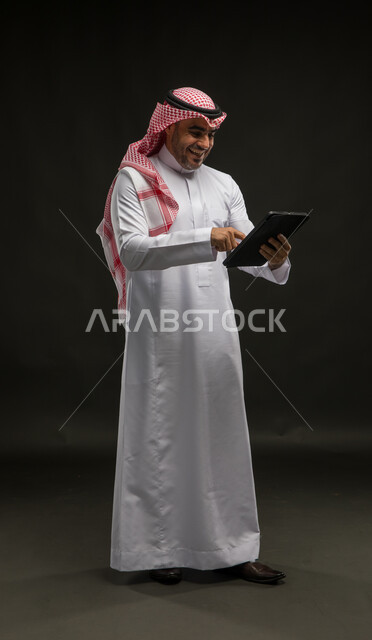 The concept of remote project management, gestures and expressions of joy and pleasure to achieve goals, use of modern technical devices, portrait of a Saudi Gulf Arab man wearing a shemagh and traditional thobe, standing upright and completing work tasks via a tablet, full-length body image, black background