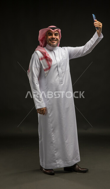 Documenting happy moments, using modern devices and technologies, a portrait of a smiling Saudi Arabian Gulf man wearing a shemagh and traditional thobe, standing straight and raising the mobile phone, looking at the camera, taking selfies, feeling happiness and joy, full body portrait, black background