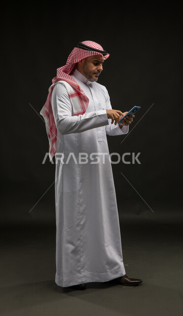 إيماءات الانزعاج وعدم الرضا, بورتريه لرجل عربي سعودي خليجي يرتدي الشماغ والثوب التقليدي يقف باستقامة ويقوم بالدردشة مع الأهل والأًصدقاء, استخدام الأجهزة والتقنيات الحديثة، الشعور بالتوتر والقلق، صورة الجسم بالكامل, خلفية سوداء
