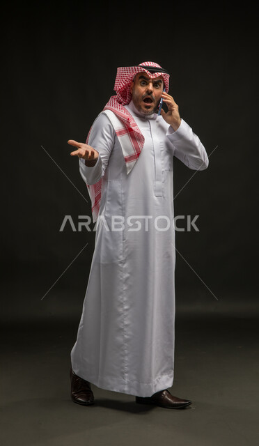 السؤال والاستفسار عن شيء ما, بورتريه لرجل عربي سعودي خليجي مبتسم يرتدي الشماغ والثوب التقليدي يقف باستقامة ويقوم بإجراء مكالمة صوتية عبر الهاتف المحمول بإيماءات الدهشة, تعابير الذهول والتفاجؤ، صورة الجسم بالكامل, خلفية سوداء