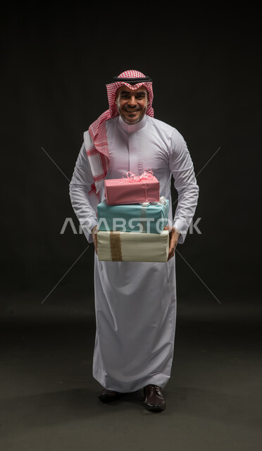 تقديم الهدايا والتهنئات، المفاجآت السعيدة في الأعياد والمناسبات، بورتريه لرجل عربي خليجي سعودي مبتسم يرتدي الشماغ والثوب التقليدي يحمل بيديه علب هدايا مغلفة ينظر الى الكاميرا بإيماءات الحماس والسرور، صورة الجسم بالكامل، خلفية سوداء