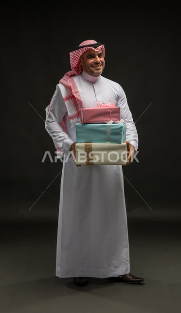 Happy surprises on holidays and occasions, a portrait of a smiling Saudi Arabian Gulf man wearing a shemagh and traditional dress, holding wrapped gift boxes in his hands, looking at the camera with gestures of enthusiasm and pleasure, offering gifts and congratulations, full body portrait, black background